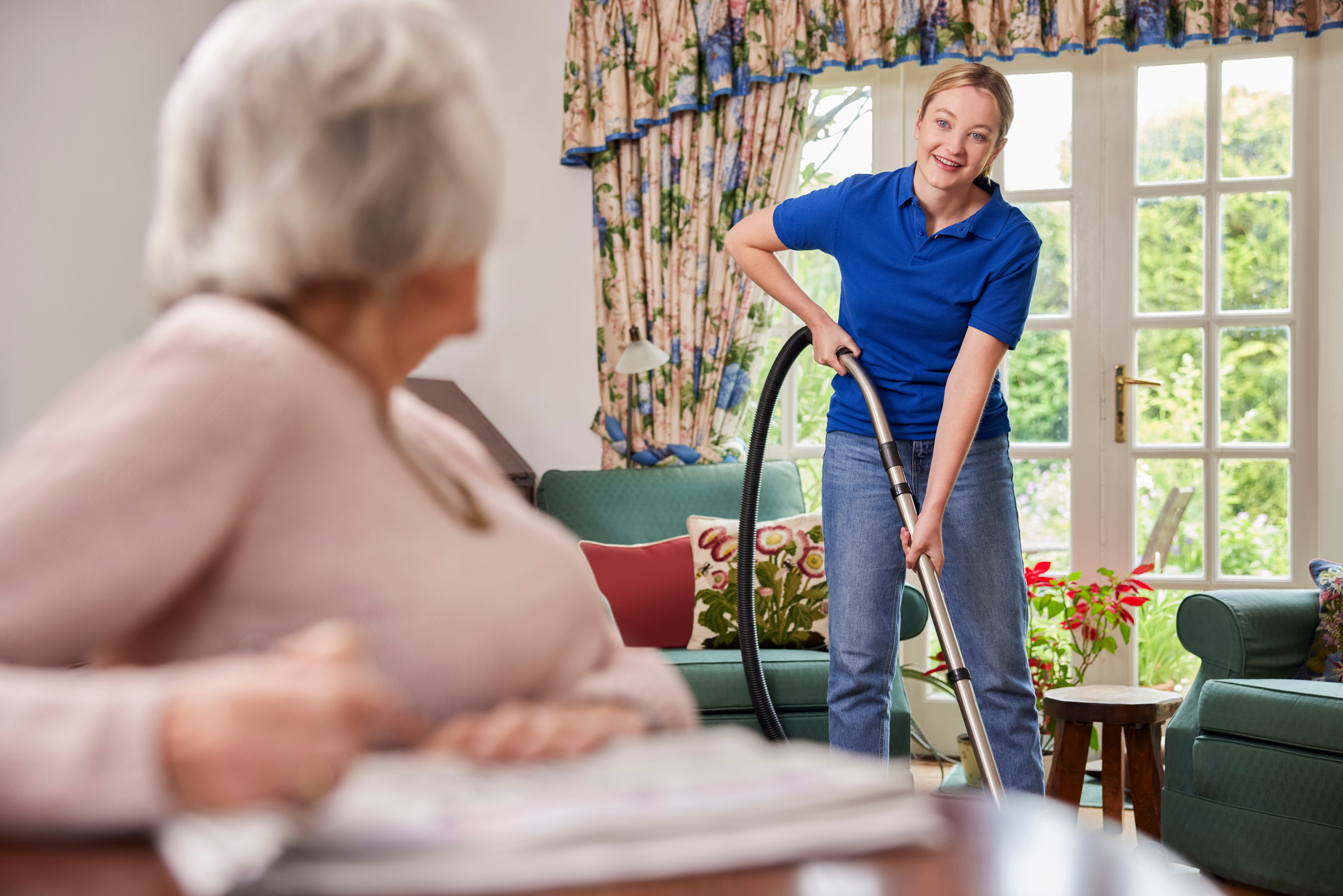 Female Home Help Cleaning House With Vacuum Cleaner And Talking To Senior Woman